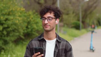 joven hermoso hombre en lentes y casual ropa cerca arriba caminando en un callejón de el ciudad parque con su teléfono inteligente y consiguiendo relajado. concepto de despreocupado urbano vida en un soleado día video