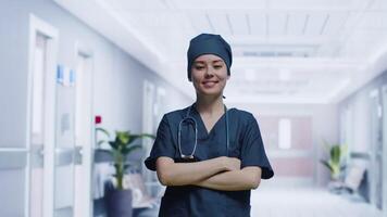 Porträt von ein jung attraktiv Frau im das Uniform von ein Arzt mit ein Stethoskop auf ihr Hals Wer Kreuzung ihr Waffen auf ihr Truhe und lächelnd im ein Krankenhaus Gang video