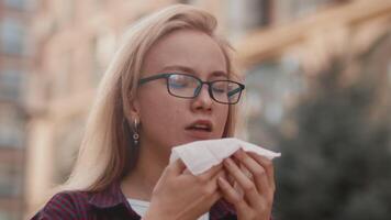 joven atractivo mujer en lentes y casual ropa estornudos dentro un pañuelo al aire libre en el ciudad calle cerca arriba. concepto de malestar y enfermedad video