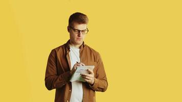 joven hermoso caucásico hombre en lentes y casual ropa en pie con un libro de copiar con un pensativo rostro, pensando y escritura en un amarillo antecedentes en un estudio video