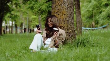 joven asiático mujer sentado debajo el árbol en el ciudad parque, Bebiendo café desde el papel taza y hablando con un amigo vía llamada en su teléfono inteligente durante el pausa en trabajo video