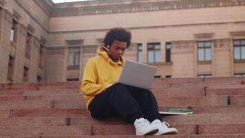 joven negro masculino programador en casual ropa sentado al aire libre en escalera y trabajando en un ordenador portátil con entusiasmo con un edificio como un fondo. concepto de flexible remoto trabajo video