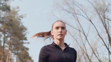 jung kaukasisch weiblich Jogger im ein schwarz Sportbekleidung Laufen mit ein zuversichtlich Gesicht auf ein Gasse im ein Herbst Stadt Park schließen hoch, Unterseite Sicht. video