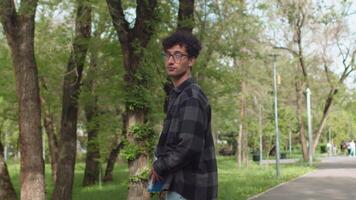 Young man in casual clothes with documents in his hands walking on an alley of the city park, then turn around and smiling. Concept of positive attitude of work video