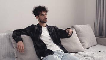 Young man in casual clothes sitting on a couch in a living room and switching channels on TV with a remote control. Concept of pleasant relaxed leisure video