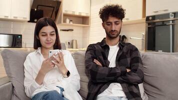 jovem ofendido chateado homem sentado em a sofá às casa Próximo para uma jovem mulher com uma Smartphone quem indiferente do dele problemas. conceito do dificuldades dentro uma casal relação video