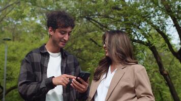 joven hombre demostración alguna cosa en un teléfono inteligente a un joven mujer, y luego ellos volverse lleno de alegría y reír, en pie en el ciudad parque durante un pausa en trabajo video