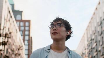 Young curly asian guy in glasses walking down a street between multi-storey buildings on a sunny day close up, looking at the sides and smiling video