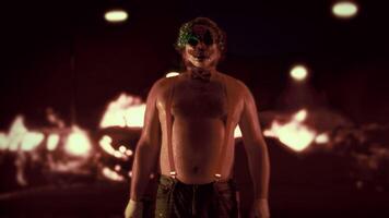 Scary man in the clown mask standing with a naked torso in suspenders and bow tie and demonstrating a chainsaw with the background of burning cars at night video