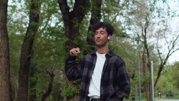 Young happy man in casual clothes walking on an alley of city park, listening music from headphones, singing along and dancing. Concept of carefree summer urban life video