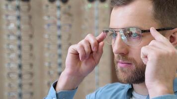 Portrait of handsome bearded guy picking new specs at optical shop, looking at mirror. Choosing glasses concept video