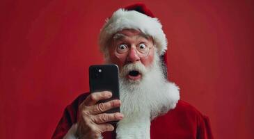 Papa Noel claus mirando a teléfono inteligente con sorprendido expresión en contra rojo antecedentes foto