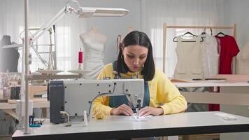 beautiful young Latin American woman is busy sewing in her creative studio, surrounded by a variety of dresses and mannequins. She is using a sewing machine to create a unique piece of clothing. Medium shot. Slow motion. 4k video