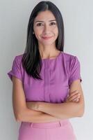 Lovely Brazilian lady in her thirties wearing a lilac blouse and a pink skirt with her arms crossed on her chest, smiling at the camera against a neutral background. photo