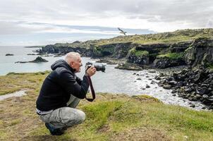 middle aged man photographer taking a picture with camera in front of a rocky coast by the sea photo