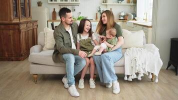 gelukkig familie Bij huis. moeder vader twee kinderen dochters ontspannende Aan sofa binnen. mam vader ouders baby meisjes kinderen kom tot rust spelen hebben pret samen. familie glimlachen lachend genieten van inschrijving moment video