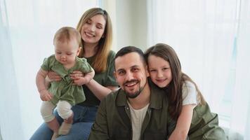 contento familia a hogar. madre padre dos niños hijas relajante interior. mamá papá padres bebé muchachas niños relajarse jugando teniendo divertido juntos. familia sonriente riendo disfrutando oferta amor momento video