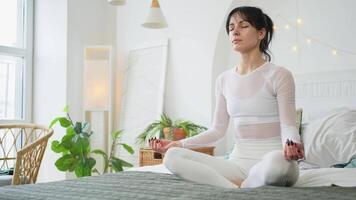 yoga opmerkzaamheid meditatie. jong gezond vrouw beoefenen yoga in slaapkamer Bij huis. vrouw zittend in lotus houding Aan bed mediteren glimlachen ontspannende binnen. meisje aan het doen ademen oefening. yoga Bij huis video