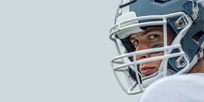 American football player in a gray helmet and white uniform with black stripes under his eyes, he stands with his left side looking to the left. White grey background. Copy space photo