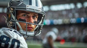 American football player wearing a gray helmet and a white uniform with black stripes under his eyes. . There is a football field and stands with fans in the background. Close-up of the face photo