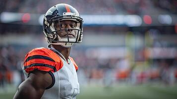 American football player in a black helmet and a white and orange uniform stands and looks to the right. On the background of the field and the stands photo