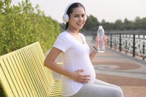 Beautiful pregnant woman drinking bottle of pure water in the park, Healthy and active pregnancy lifestyle concept. photo