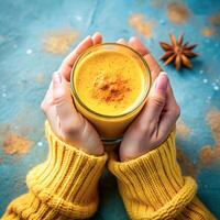 Hands holding glass of organic curcumin honey golden milk, Indian turmeric latte on blue background. Top view spices yellow chai natural drink healthy food concept photo