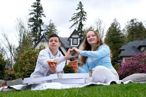 adolescente estudiantes chico y un niña en un almuerzo descanso a escuela. selectivo atención joven Adolescente Pareja en blanco camisas teniendo picnic en girasol campo en puesta de sol. comiendo Pizza y Bebiendo champán foto