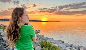 a teenage girl in a Green T-shirt shoots a beautiful sunset on the Pacific Ocean she holds an iPhone 13 phone in her hands blond red hair visible stones setting sun sea and sky photo