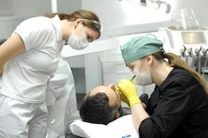 dental clínica médico elimina diente paciente en amarillo guantes asistente mira a estudiante estudiando último tecnología hembra médico y paciente hombre relleno caries podrido diente prótesis cepillado dientes foto