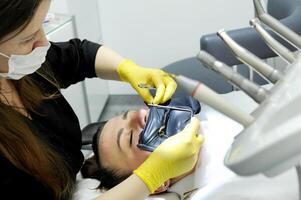 terraplén abrazadera para terraplén el proceso de dental prótesis utilizando un microscopio, hembra médico en amarillo guantes, paciente dentista con paciente, utilizando dental curación ligero foto