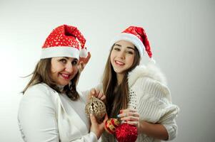 mamá y hija son vestido en blanco con rojo nuevo años sombreros en su cabezas participación Navidad juguetes muchos emociones de alegría en un blanco antecedentes fiesta invitación hogar celebracion foto