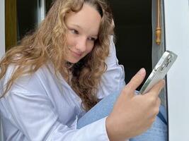 a girl in a white shirt a teenager sits on a white windowsill the view from the street she can be seen in the window in her hands she holds a white iPhone 13 pro max she looks at the screen calmly photo