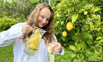 it's just a lemon paradise, a teenage girl stands against the backdrop of a lemon tree with lemons and drinks lemonade from a glass glass in her other hand, she holds a jug of drink photo