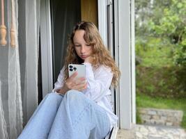 a girl in a white shirt a teenager sits on a white windowsill the view from the street she can be seen in the window in her hands she holds a white iPhone 13 pro max she looks at the screen calmly photo