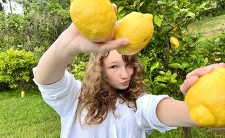 a fair-haired girl juggles lemons against the backdrop of a lemon tree in a tropical country, she is dressed in a white shirt she laughs and has fun photo