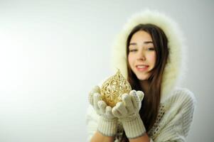 tarjeta postal con un hermosa mujer nieve doncella joven hermosa niña en mitones sostiene en su manos un juguete ese es para el Navidad árbol ella es vistiendo un capucha con piel blanco guantes sostener un dorado pelota foto