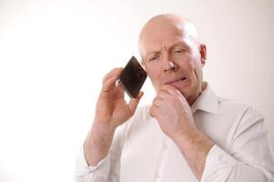 Senior man feeling upset having phone conversation depressed by hearing bad news adult man in white shirt grabbed his head and is upset, he listens to the news from the phone, he is talking to someone photo