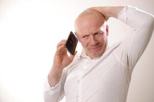 Senior man feeling upset having phone conversation depressed by hearing bad news adult man in white shirt grabbed his head and is upset, he listens to the news from the phone, he is talking to someone photo