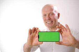 Portrait of positive funky aged person indicate finger empty space proposition shows off something on smartphone with green screen to camera. Focusing on smartphone with chromakey photo