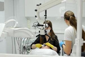 un joven niña en un dental silla y un dentista quien se sienta siguiente a a él. él mira a el dientes con un dental microscopio y sostiene un dental rebaba y un espejo en su manos. dentista y paciente durante procedimiento foto