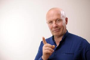 Emotions of an adult handsome bald man A man on a white background in a blue shirt The man smiles slyly and looks into the frame. He shows a thumbs up as if for some reason he wants to draw attention. photo