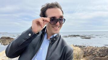 A handsome French man in black sunglasses stands on the shores of the Atlantic Ocean, he has black hair, a leather jacket and he flies with his hands to the sides travel advertising photo