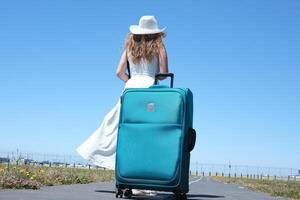 young girl in white dress and huge hat with a suitcase travels around globe Close up woman legs walks on asphalt road and traveling world with travel suitcase . Female hitchhiking around country photo