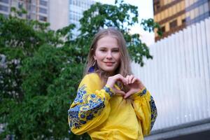 cute beautiful blonde girl young woman looks into the frame and makes a heart with her hands joyful in a yellow shirt on a white-green background street love peace in Ukraine joy calmness photo