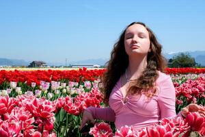 un hermosa joven niña elevado su cara en el viento mira cerrado su ojos inhala Fresco aire soportes en un campo de tulipanes rojo tulipanes con un blanco frontera en contra claro azul cielo frescura alegría libertad será foto