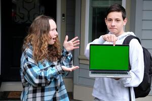 two teenagers in frame boy and girl friend points finger at green chroma key screen on computer laptop girl is surprised with fingers spread open mouth surprised schoolchildren on street advertising photo
