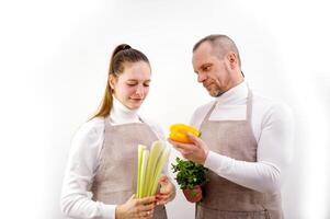 marido esposa en cocina delantales Mira a vegetales gente ropa y antecedentes son blanco ellos tener pimientos su manos apio perejil cilantro ellos son vegetarianos Fresco vegetales cocinar cena foto