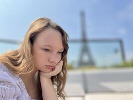 the face of a beautiful girl she is sad leaning on her arm in the distance you can see the Eiffel Tower the girl is young she is a teenager photo