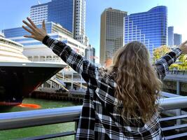 un alegre adolescente niña soportes en contra el fondo de crucero Embarcacion Canadá ciudad Vancouver en un hermosa vestido, ella elevado su mano, usted lata anunciar un viaje agencia viaje a Vancouver antes de Cristo foto